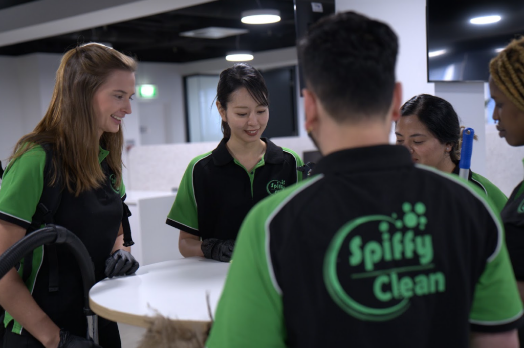 Team of Spiffy Clean staff in green and black uniforms gathered around a round white table, smiling and talking.