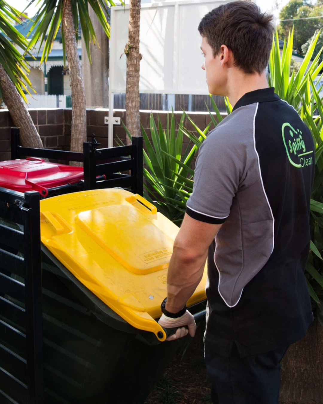 Picture1342 | Spiffy Clean - Commercial & Office Cleaning Services | Spiffy Clean - Commercial & Office Cleaning Services Maintenance worker in a gray and black uniform labeled 'Spiffy Clean' places a yellow recycling bin into a black outdoor bin.