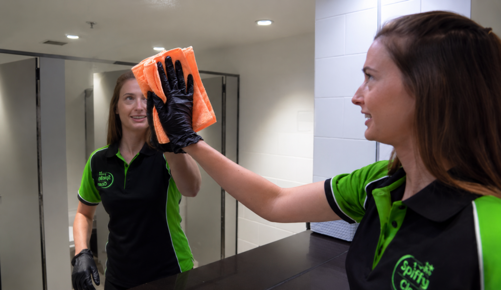 Two cleaners in green and black uniforms high-five across a dark counter; one wears black gloves and holds an orange microfiber cloth in a bright restroom.